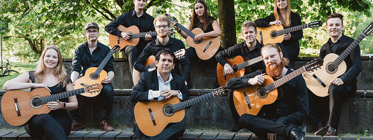 Ein Gruppenfoto des Gitarrenensembles. Die Zehn Musiker:innen halten ihre Gitarren auf dem Schoß.